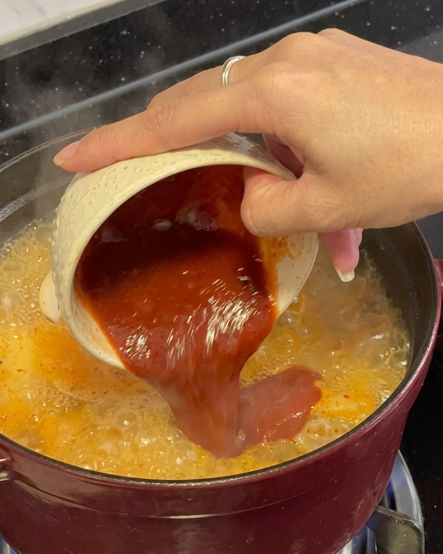 A thick, dark red chili seasoning mixture being poured from a small speckled bowl directly into a bubbling red seafood stew.