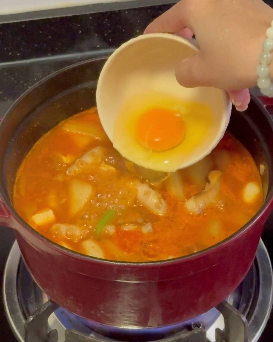 A raw egg with a bright yellow yolk being gently poured from a small bowl into a vigorously boiling red seafood stew.
