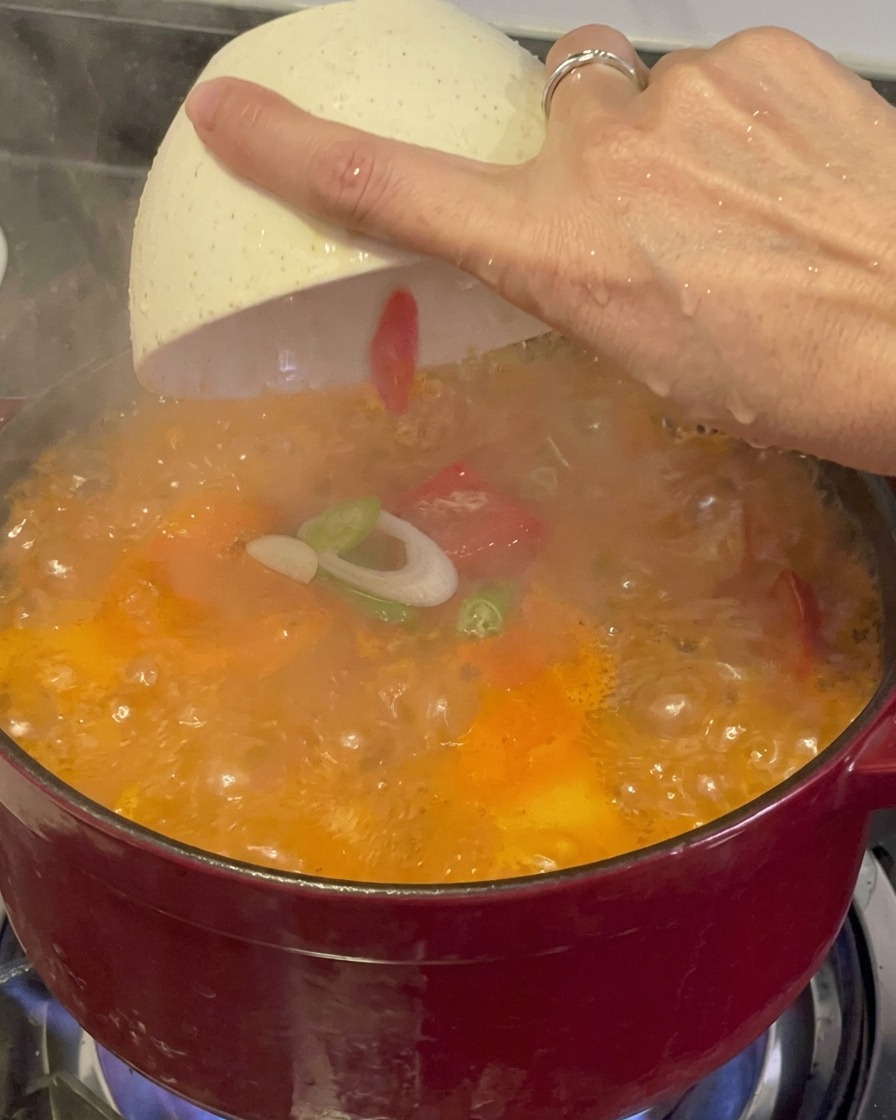 Freshly sliced green scallions and red chilies being sprinkled from a white bowl over a steaming, bubbling red kimchi stew.