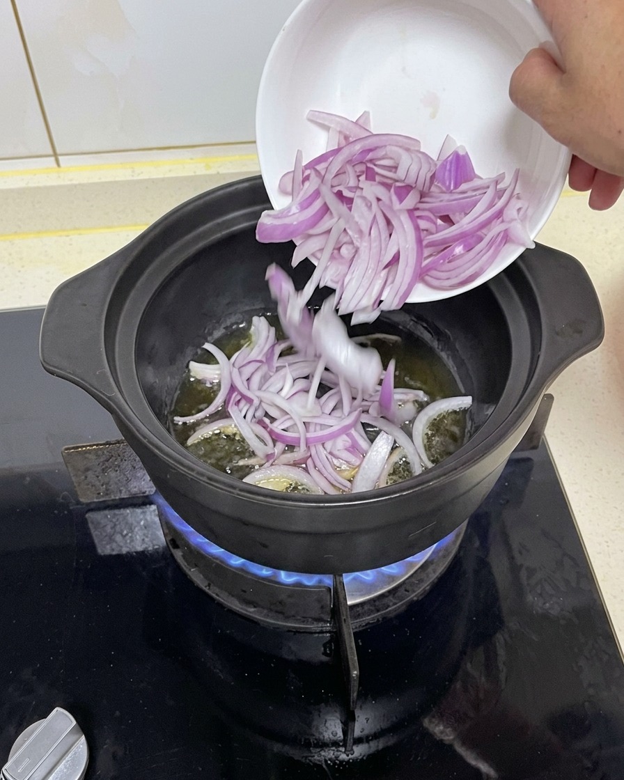 Slices of red onion being transferred from a small white bowl into a hot black cooking pot with oil over a blue gas flame.