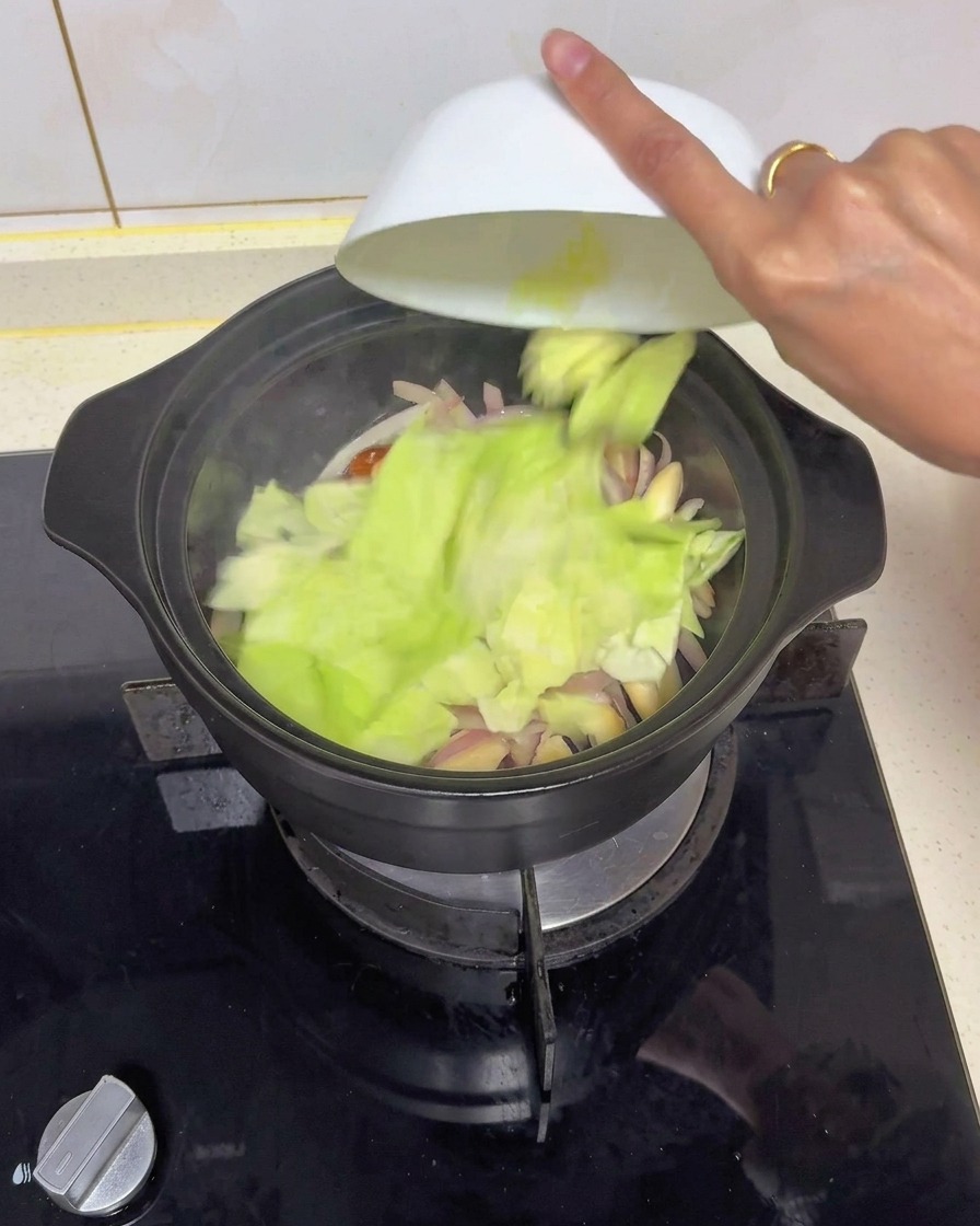 Chopped pieces of fresh green cabbage being emptied from a white bowl into a black pot filled with sautéed onions and mushrooms.