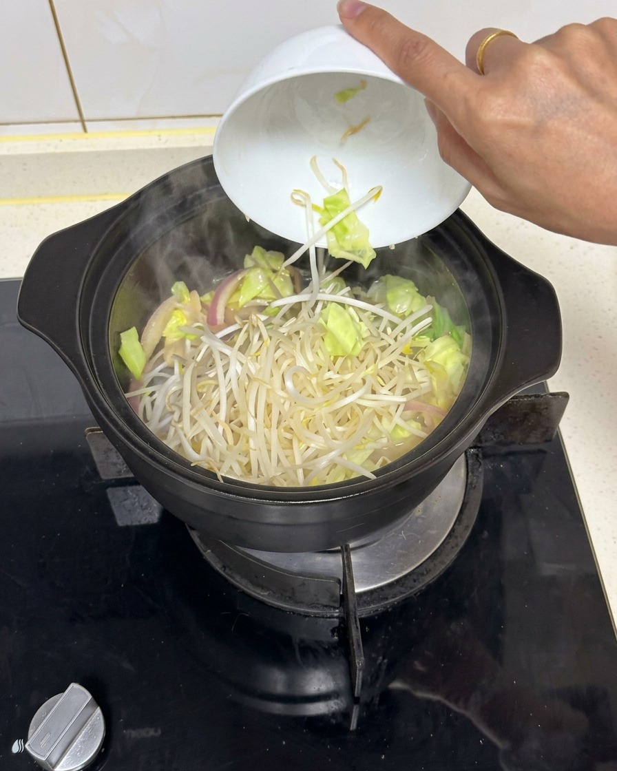 A heap of fresh bean sprouts being dropped from a white ceramic bowl into a steaming black pot of mixed stir-fried vegetables.