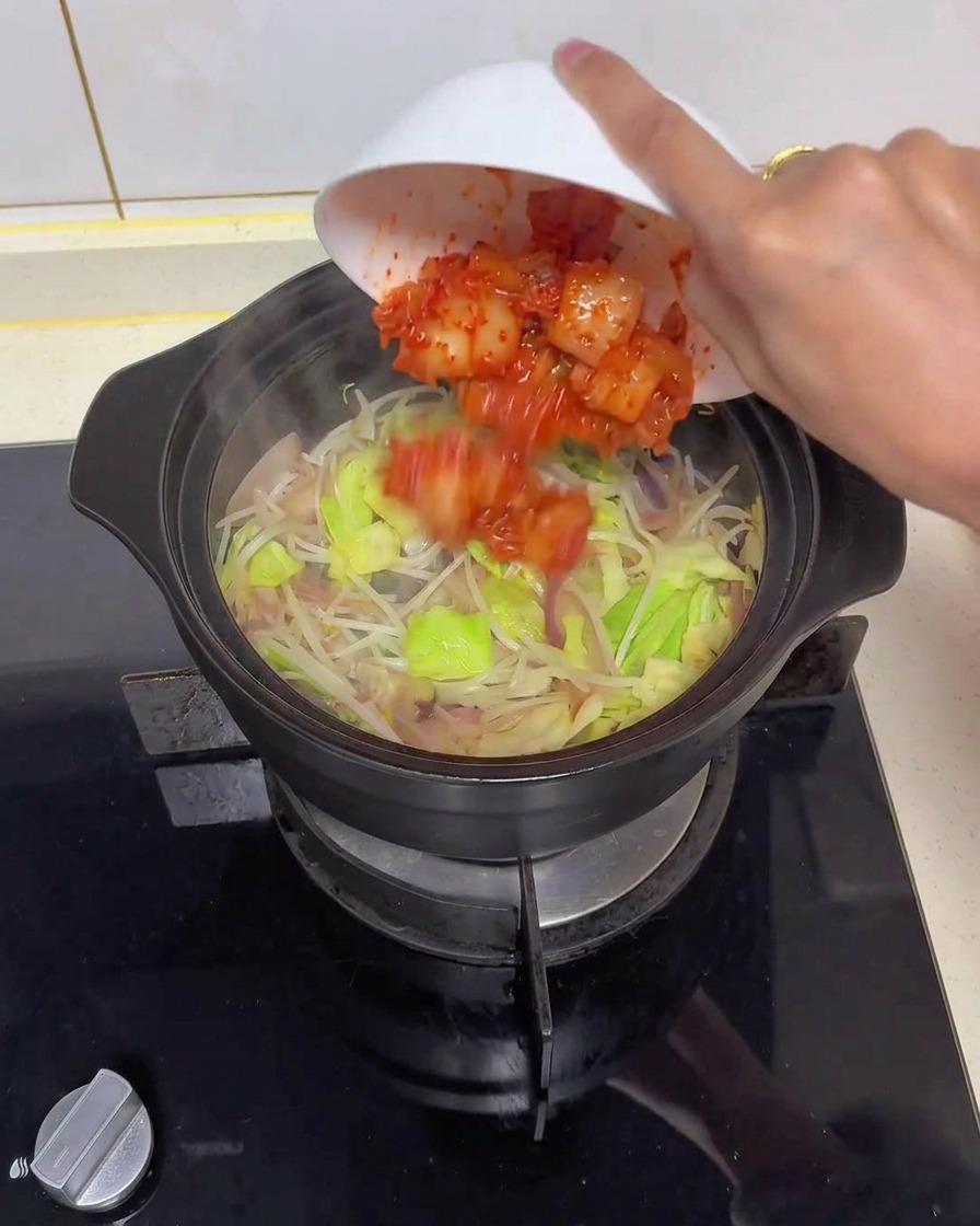 A hand tipping a white bowl to add bright red chopped kimchi into a black ceramic cooking pot filled with translucent onions, green cabbage, and bean sprouts.