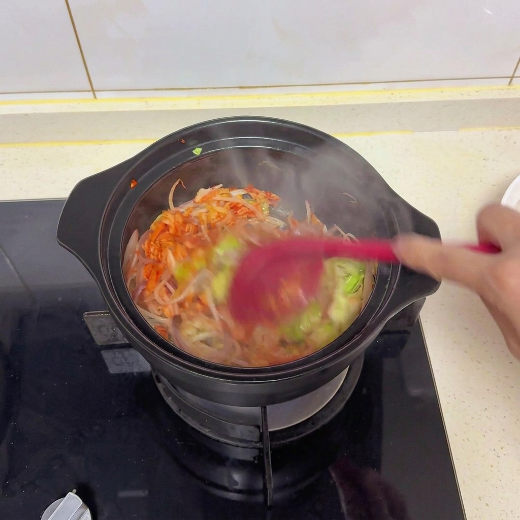 Stir-frying mixed vegetables and kimchi in a black pot with a red spatula to mix in the salt, soy sauce, and seasonings.