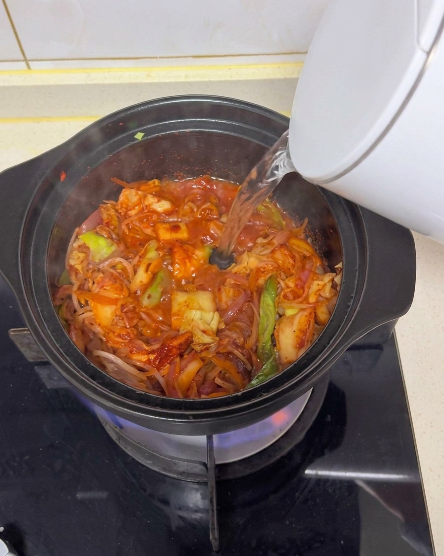 Hot water being poured from a white electric kettle into a black clay pot on a gas stove, mixing with stir-fried ingredients to form a red broth.