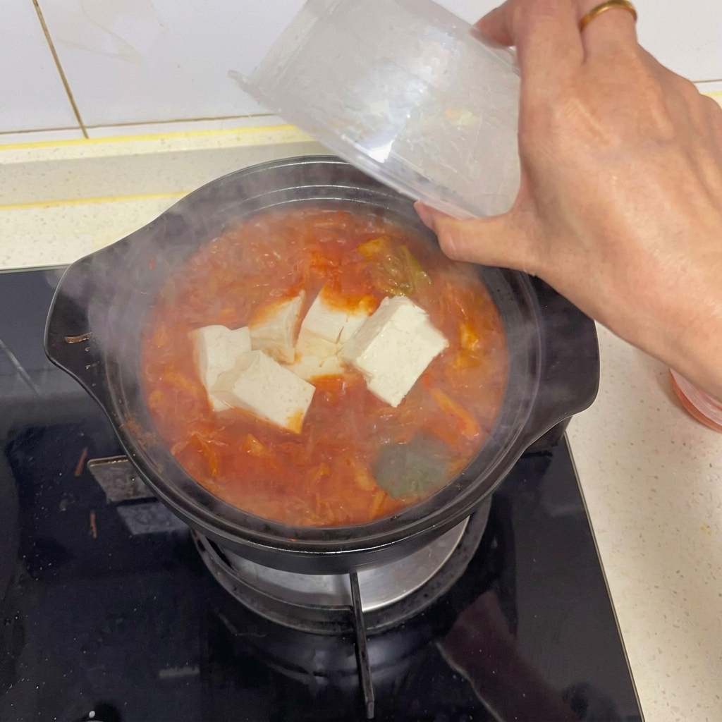 A hand gently adding large cubes of white tofu from a clear plastic container into a boiling pot of red kimchi soup.