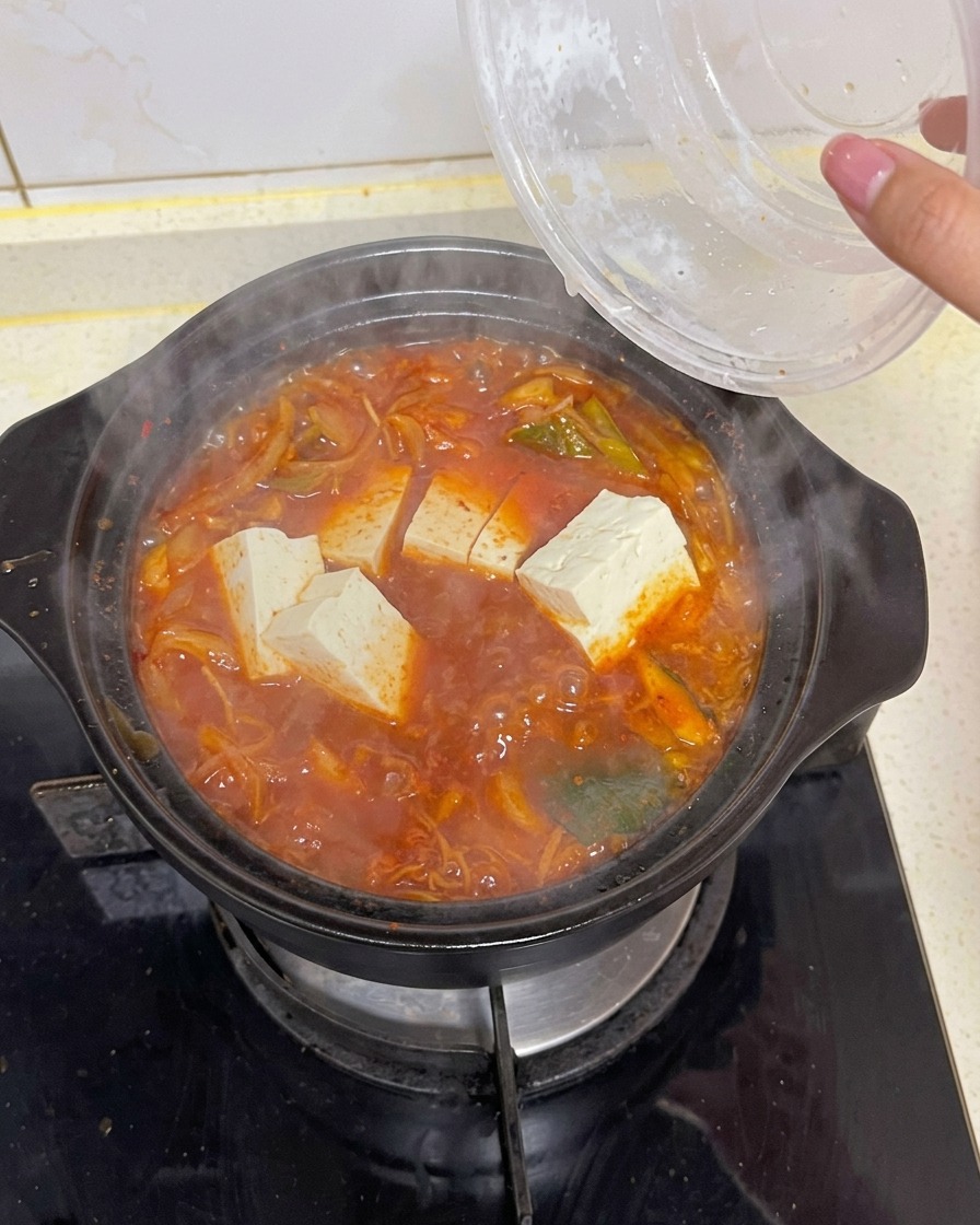 Slabs of clean white rectangular tofu being dropped from a clear container into a bubbling pot of dark red kimchi soup containing greens and kelp.