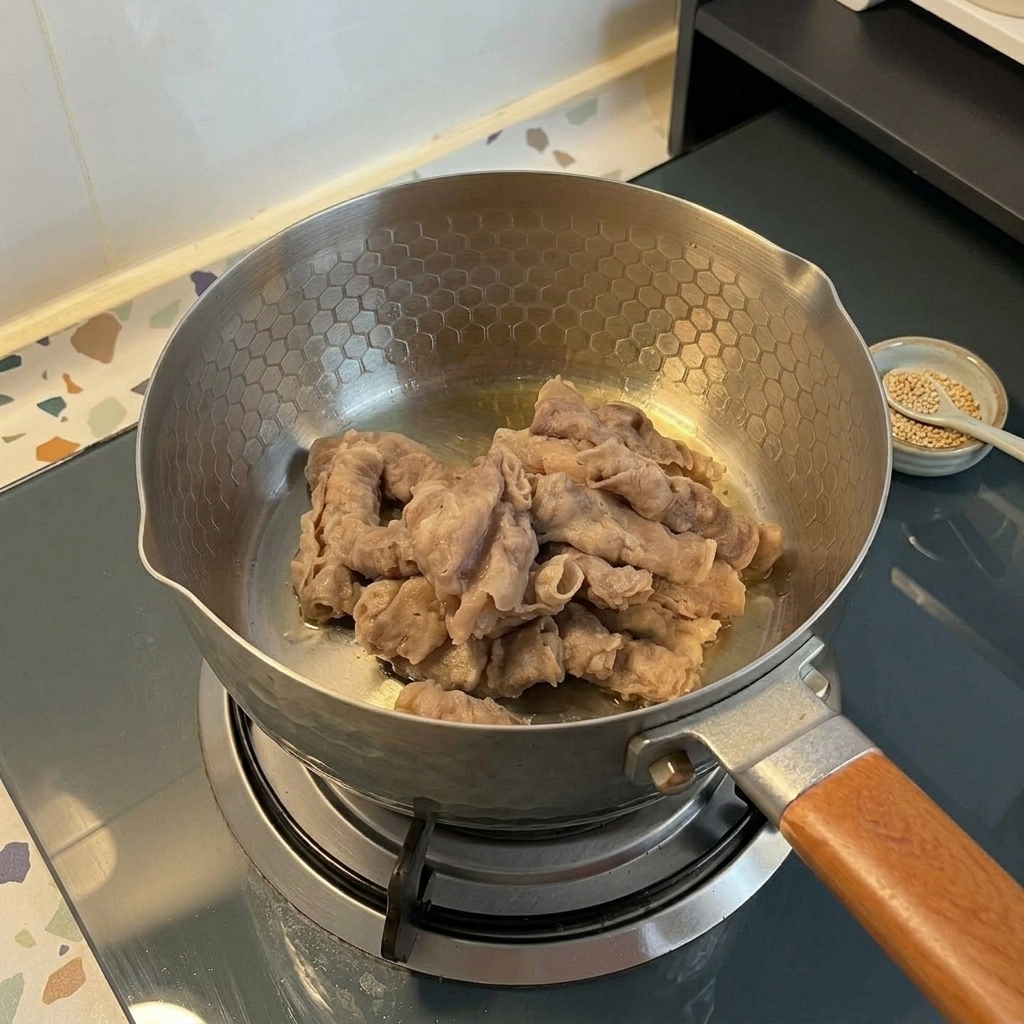 Thinly sliced beef being stir-fried in a stainless steel pot with a honeycomb pattern on a gas stove.
