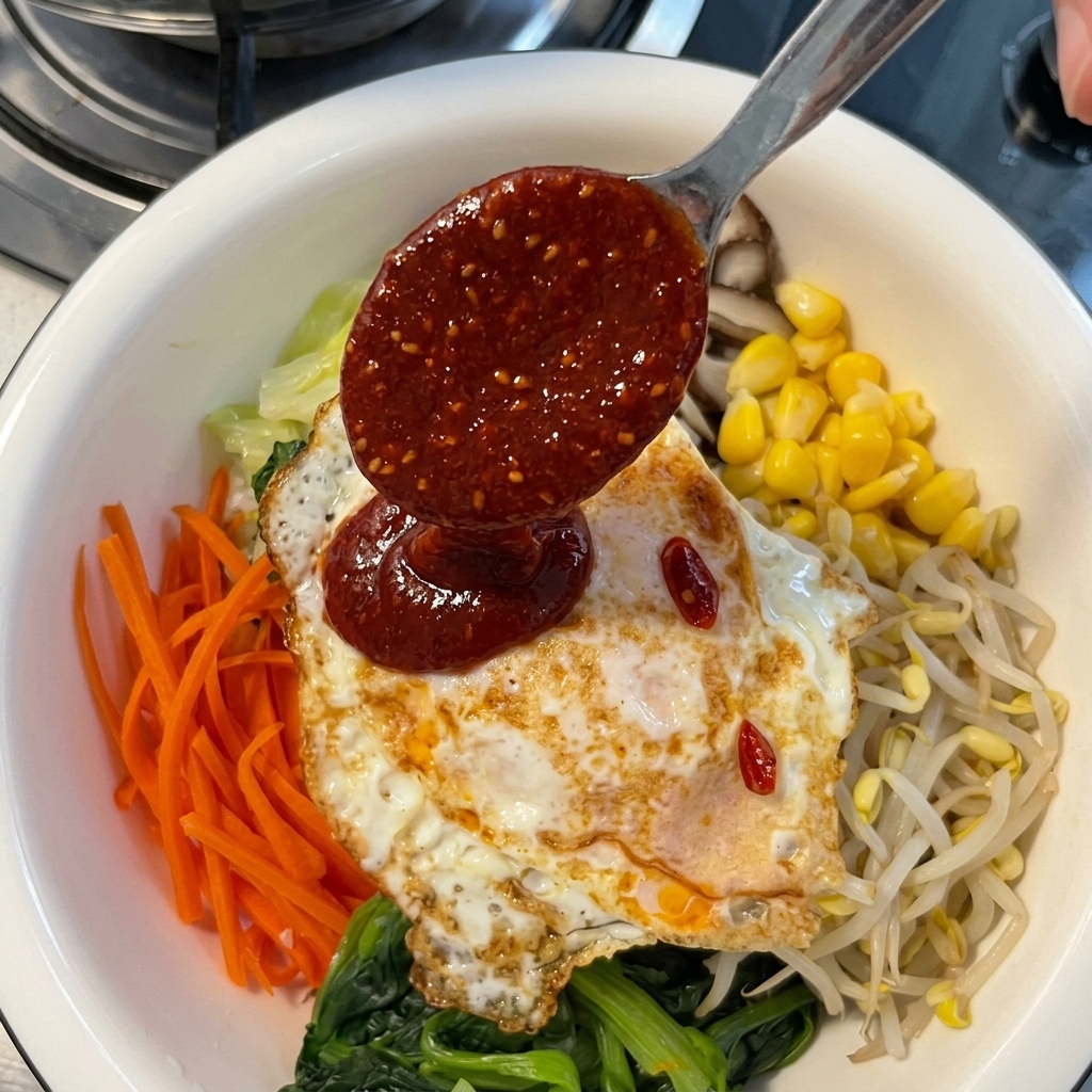 Drizzling a thick red chili paste sauce from a metal spoon onto a fried egg layered over a colorful assortment of bibimbap vegetables.