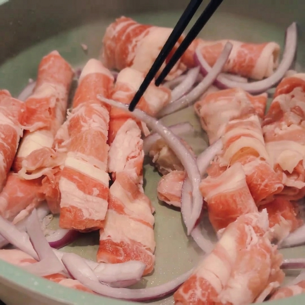 Chopsticks stirring thin slices of semi-raw beef and red onions in a light green cooking pan.