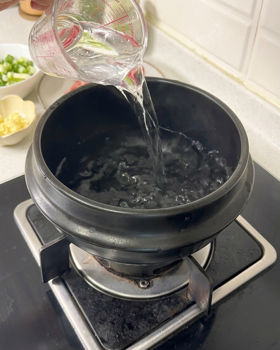 Water being poured from a clear measuring cup into a black earthenware pot on a gas stove burner.
