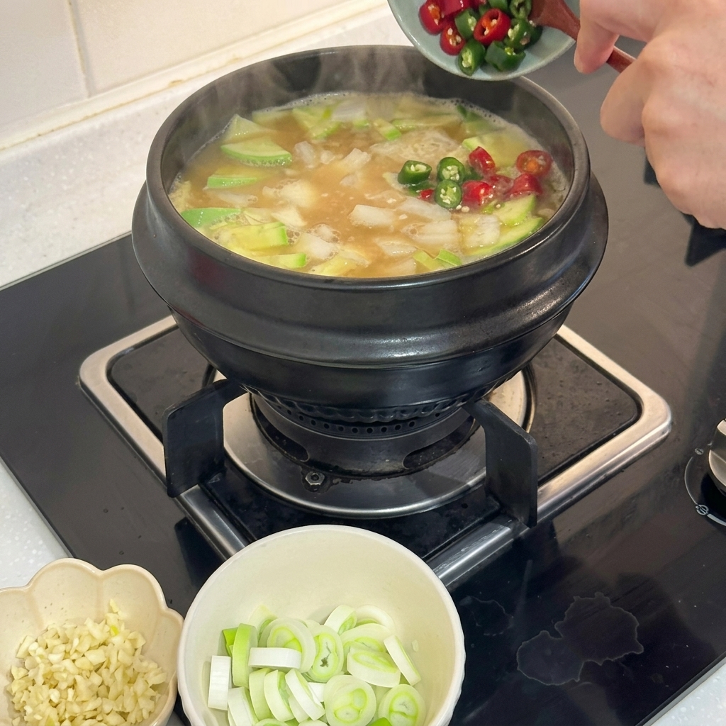Finely chopped red and green chili peppers being slid into a bubbling pot of soybean paste stew.
