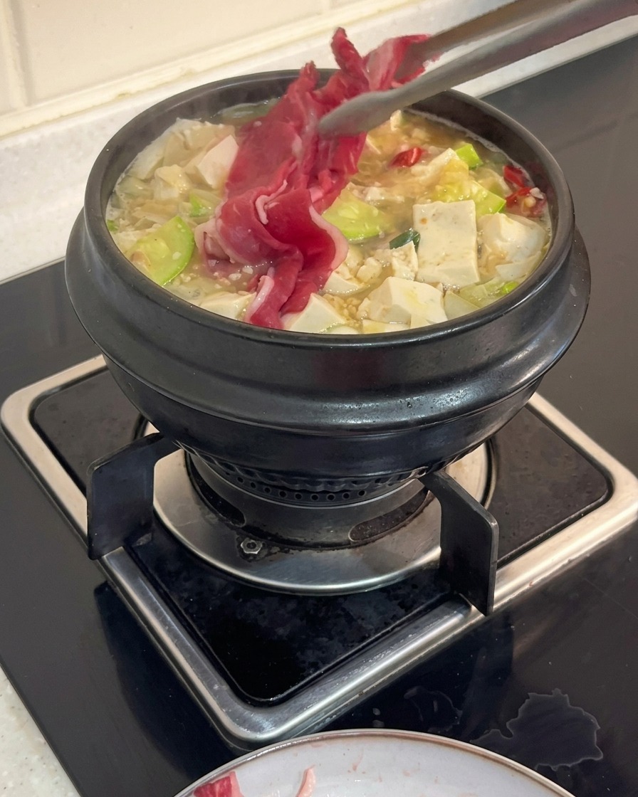 Kitchen tongs placing thin slices of raw beef brisket into a boiling pot of Korean stew filled with tofu and zucchini.