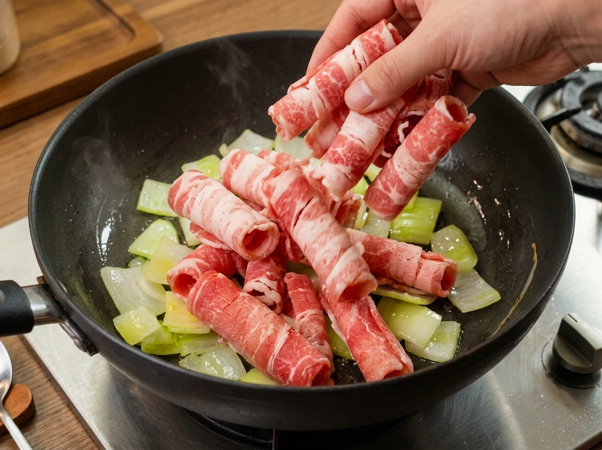 Rolled slices of raw beef being placed into a pan with translucent sautéed onions.