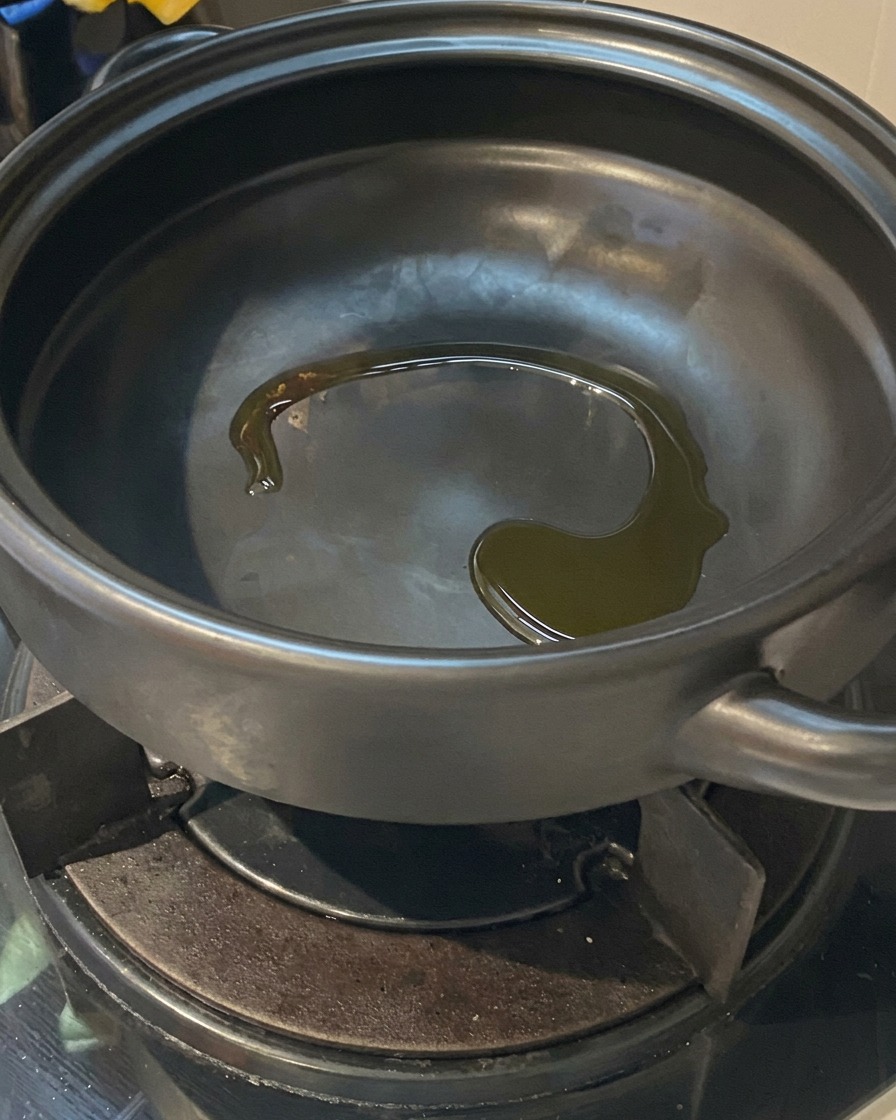 Cooking oil glistening at the bottom of a heated black clay pot sitting on a gas stove burner.