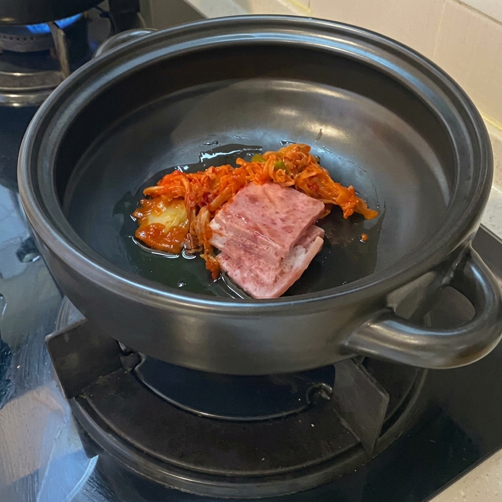 Slices of raw pork belly and vibrant chopped kimchi resting in oil at the bottom of a dark ceramic clay pot.