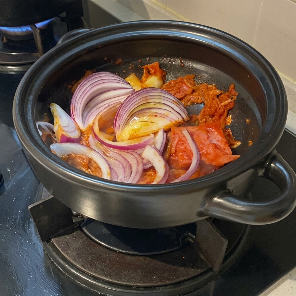 Thick slices of vibrant red onion layered over a seasoned stir-fry of kimchi and pork belly inside a clay cooking pot.