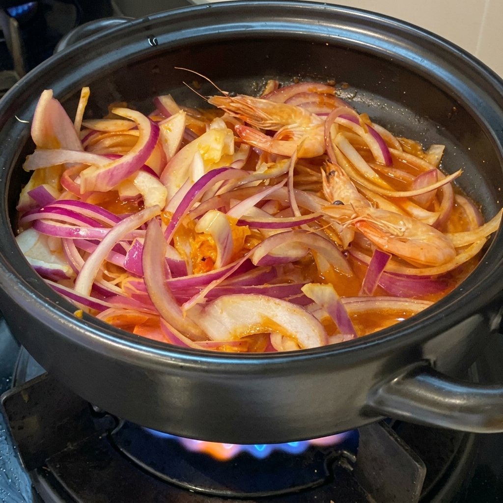 Fresh whole pink shrimps nestled among sliced red onions and kimchi in a black clay pot on a lit gas stove.