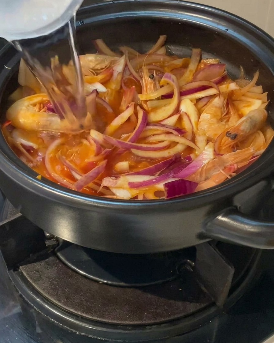 A stream of clear water being poured from a cup into a clay pot filled with sliced red onions and whole shrimp.