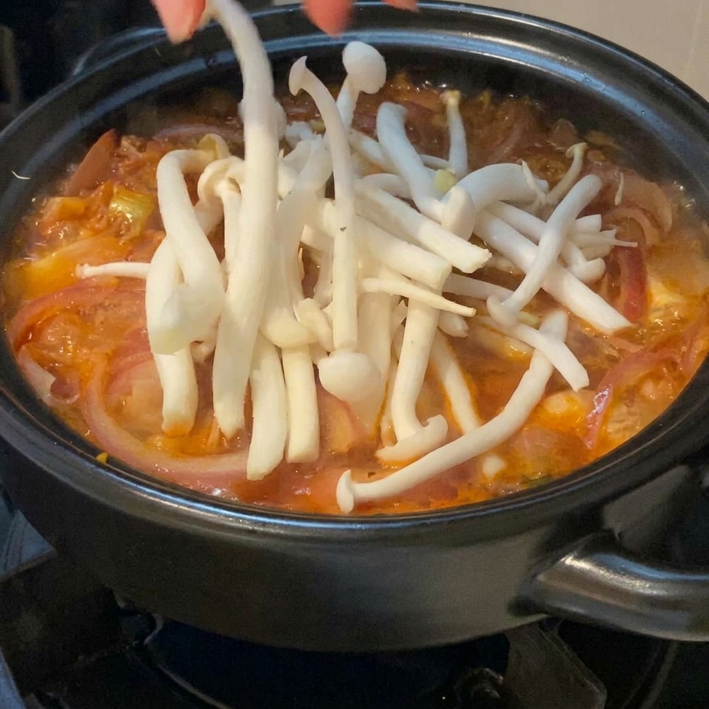 Fresh white seafood mushrooms being carefully layered by hand onto a bubbling red kimchi soup inside a black clay pot.