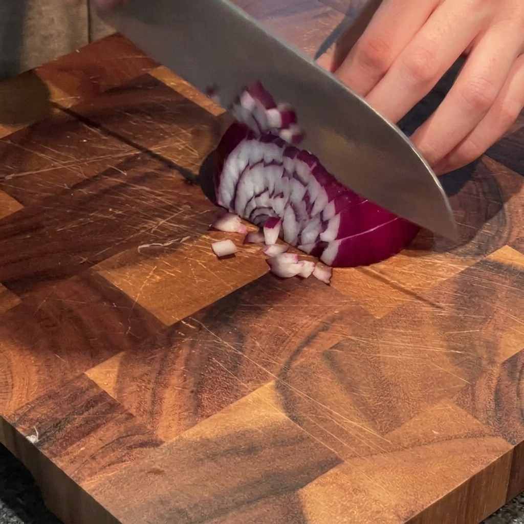 A hand using a chefs knife to dice a red onion into small pieces on a wooden cutting board.