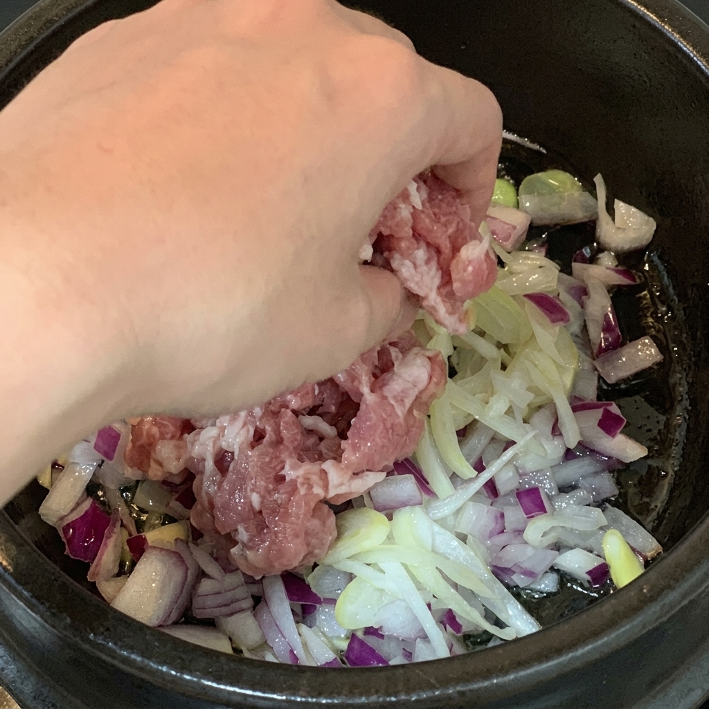 Raw pork belly pieces being added into a cooking pot filled with diced red onions and shredded scallions.