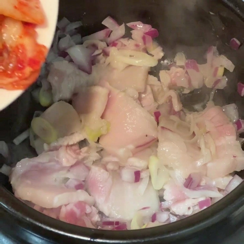 Chopped kimchi cabbage being added from a plate into a cooking pot containing pork belly pieces and red onions.