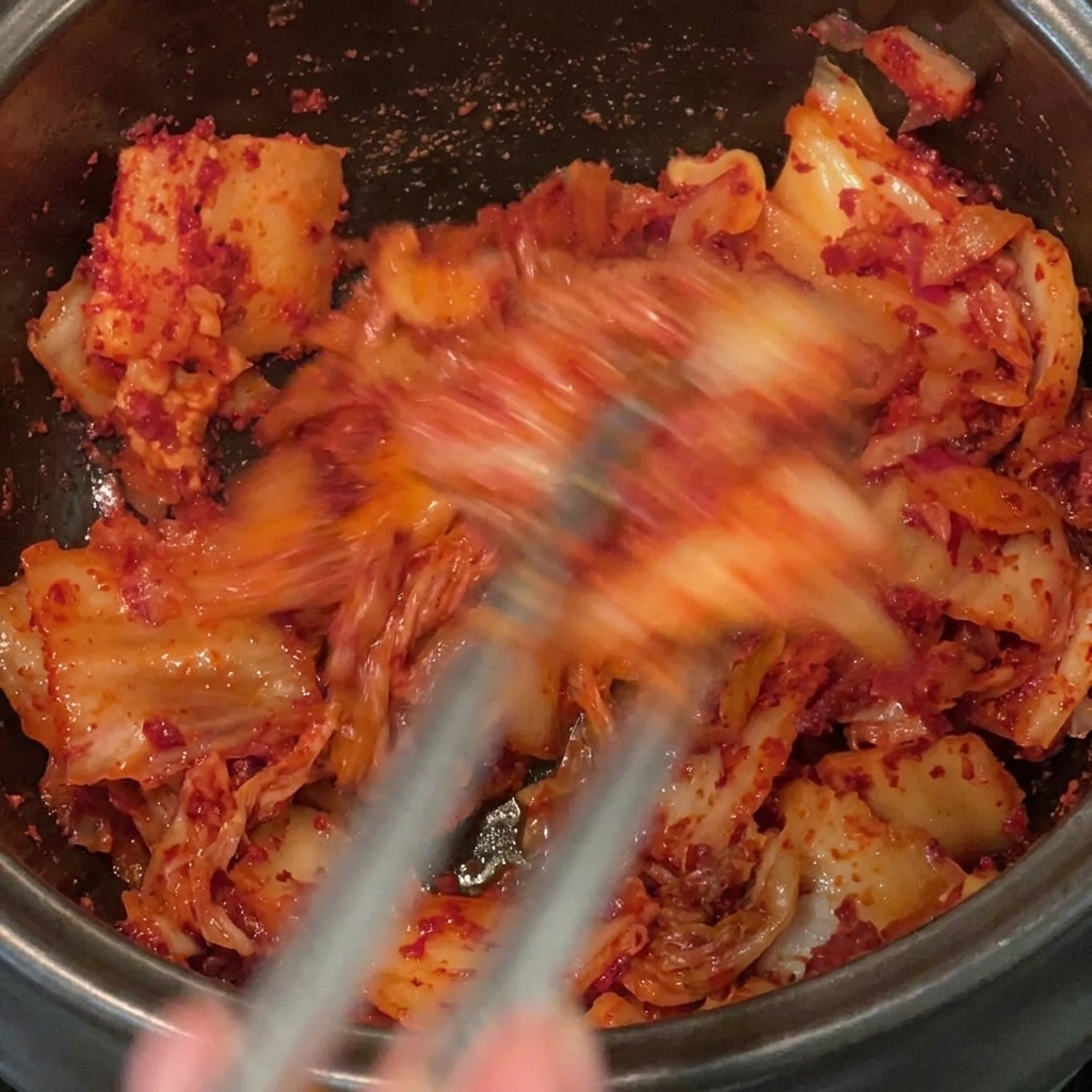 Stirring and thoroughly mixing red chili powder into chopped kimchi and pork belly inside a black pot.