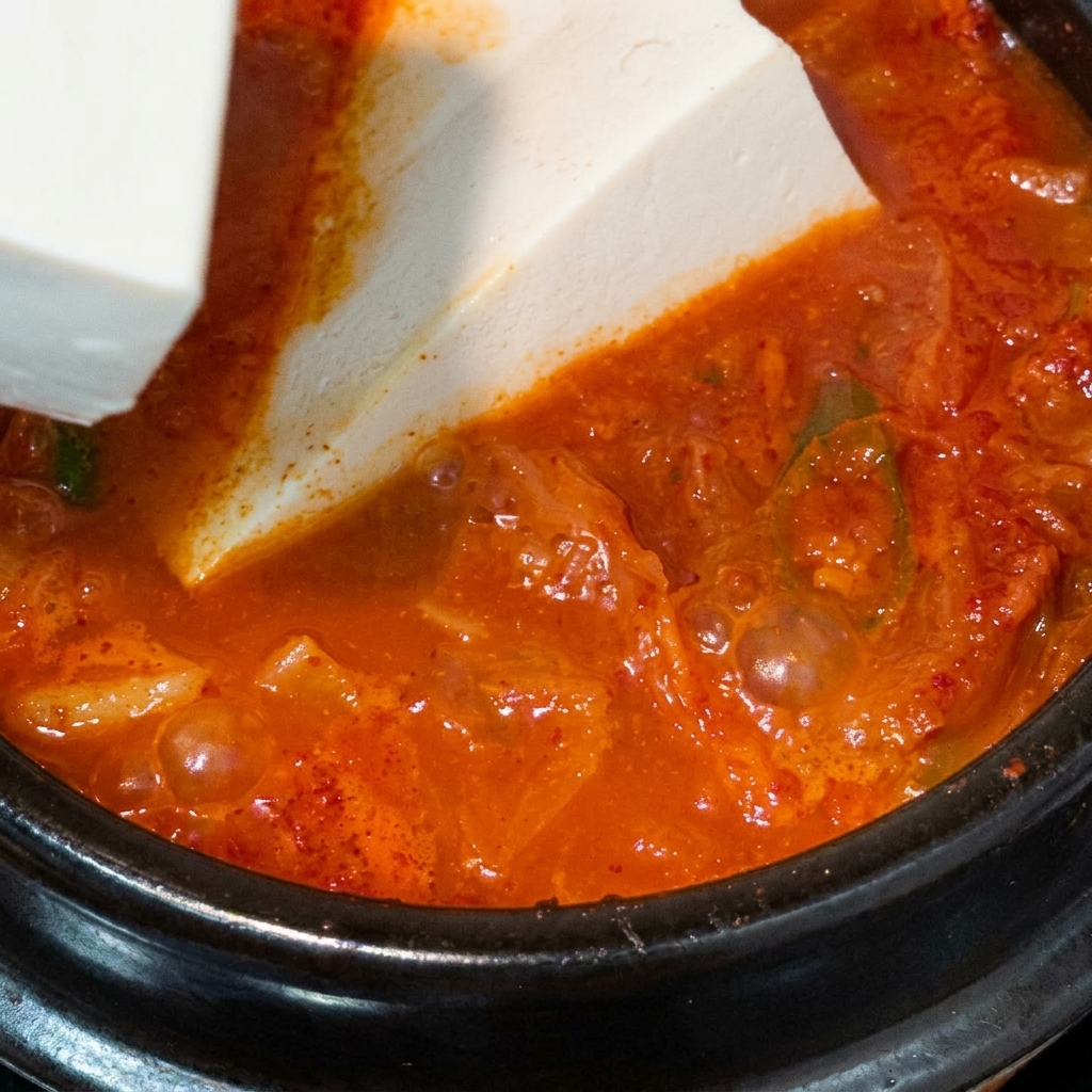 A large block of white silken tofu being carefully placed into a boiling pot of red kimchi soup.