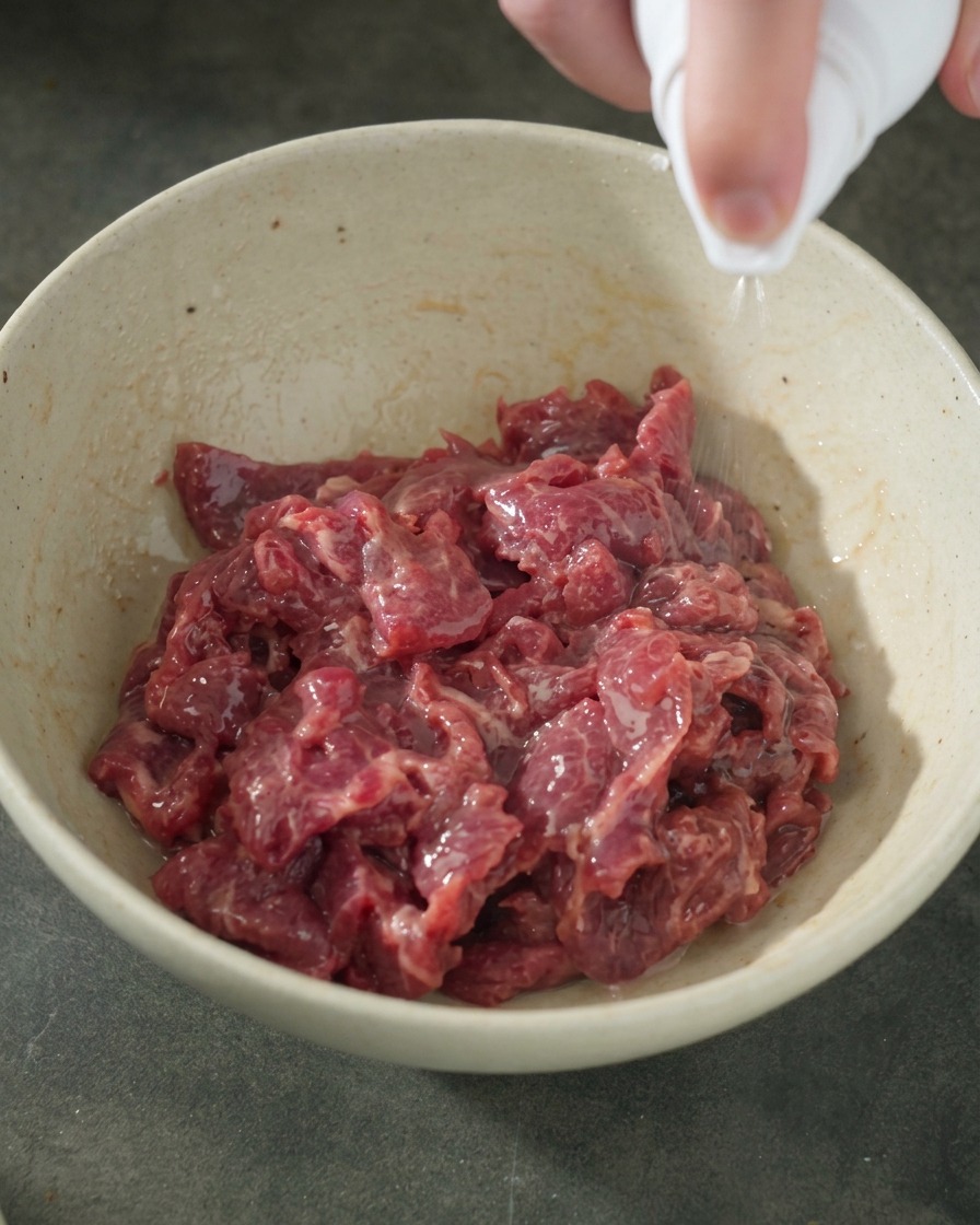 A hand holding a white spray bottle, spraying a fine mist of cooking oil onto a bowl of well-marinated, glossy raw beef slices.