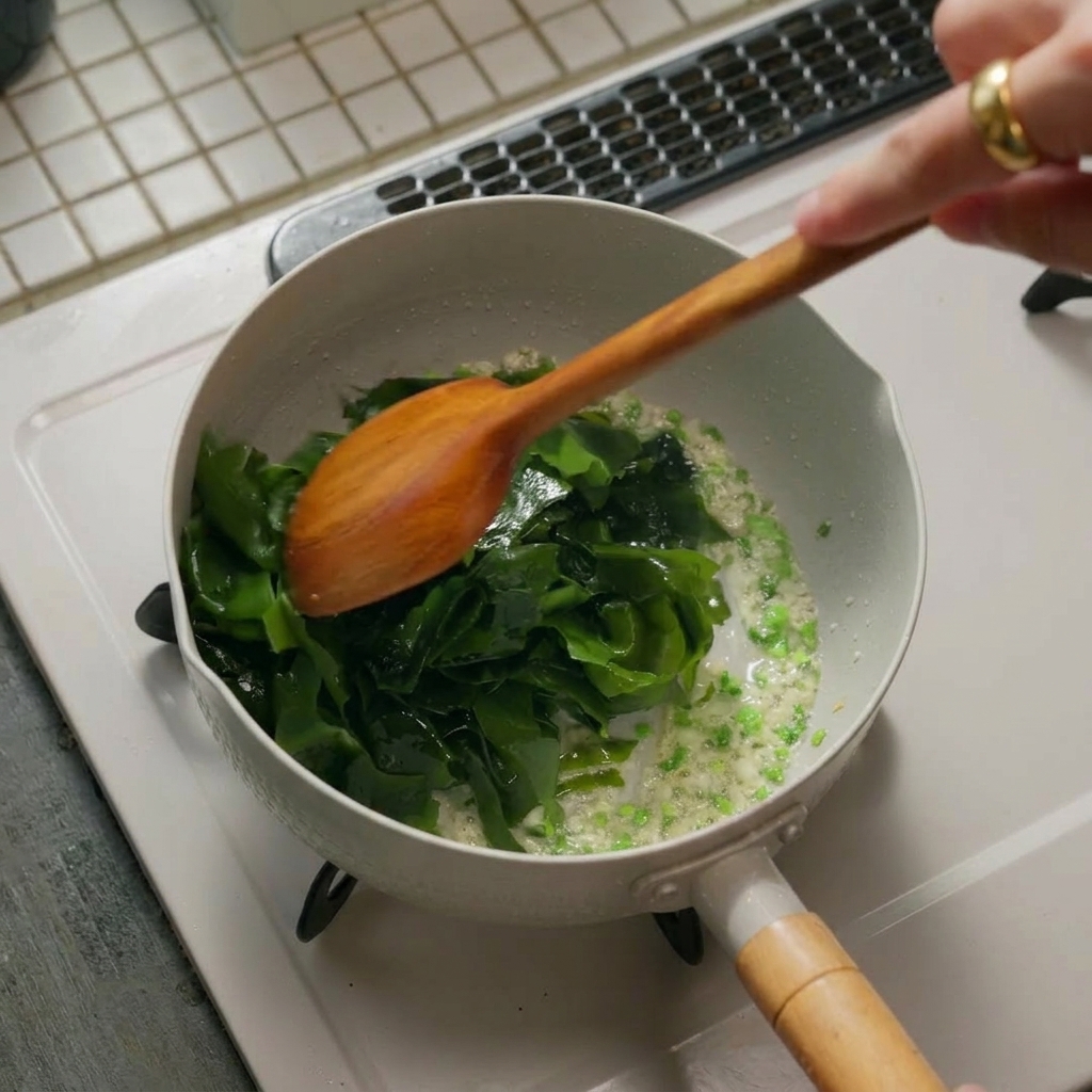 A pile of vibrant green rehydrated seaweed added into a white pan with sautéed aromatics, being stirred by a wooden spatula.