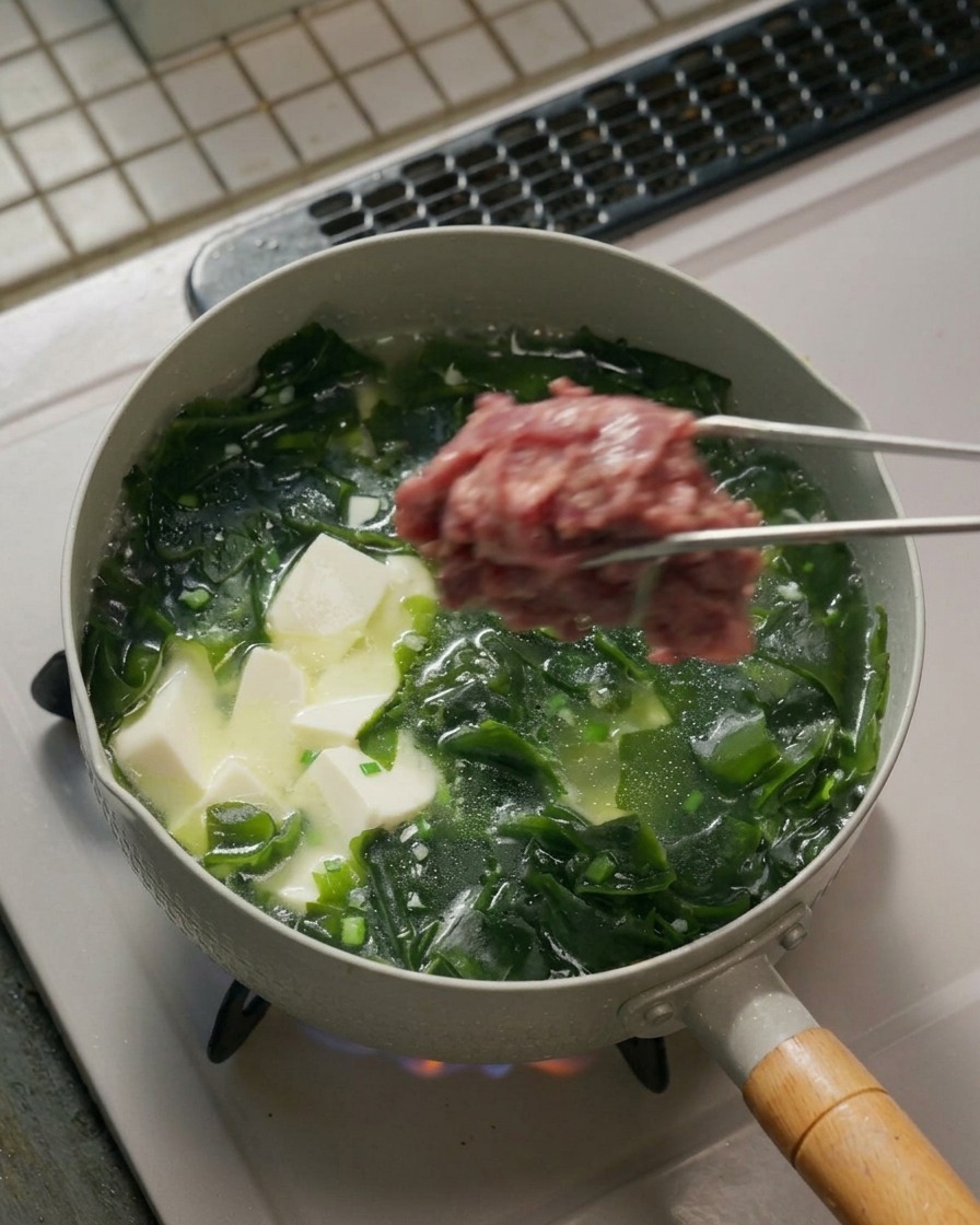 Metal tongs holding a clump of raw marinated beef slices over a boiling pot filled with seaweed and tofu soup.