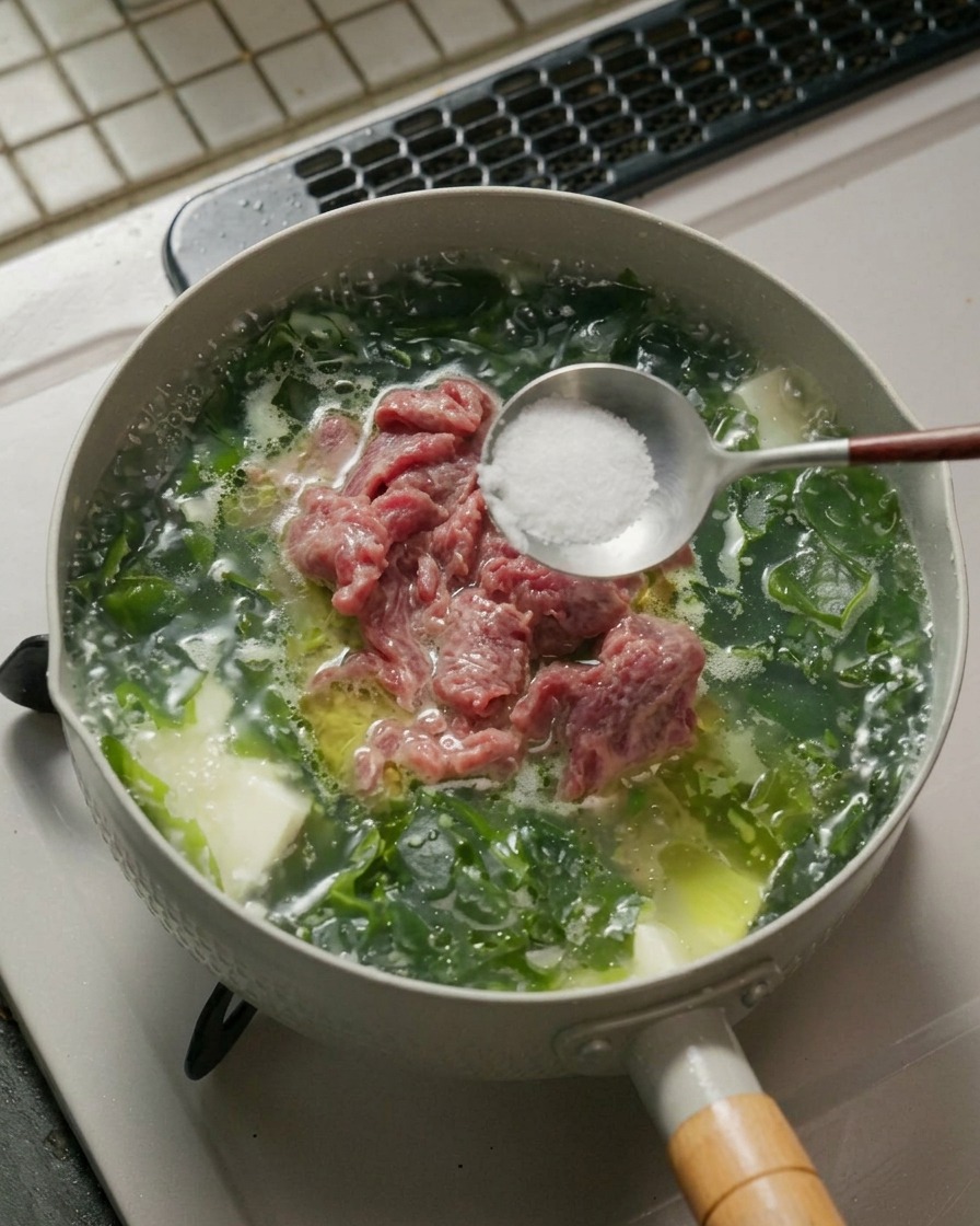 A metal spoon holding white salt and pepper powder over a pot of simmering seaweed beef tofu soup.