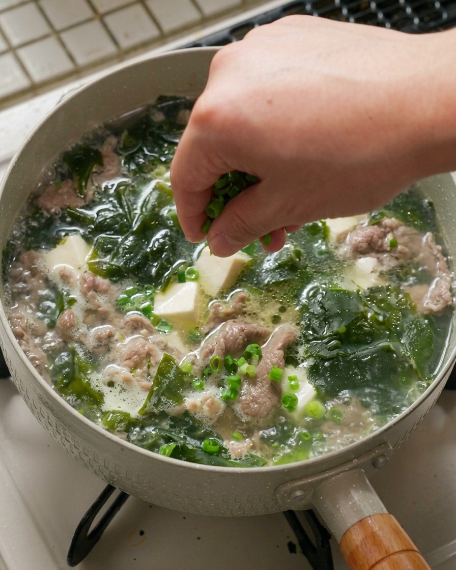 A hand sprinkling a generous pinch of finely chopped green scallions over a hot, simmering pan of seaweed soup containing white tofu cubes and pieces of cooked beef.