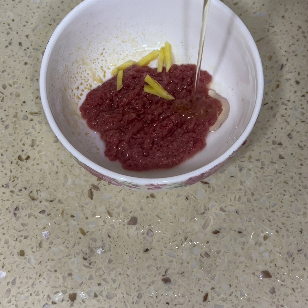 Minced raw beef in a white ceramic bowl topped with julienned ginger slices, with a thin stream of baby cooking oil being poured over it.