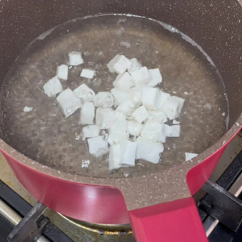 Small cubes of soft white tofu swirling and blanching in boiling water inside a pink non-stick cooking pot.