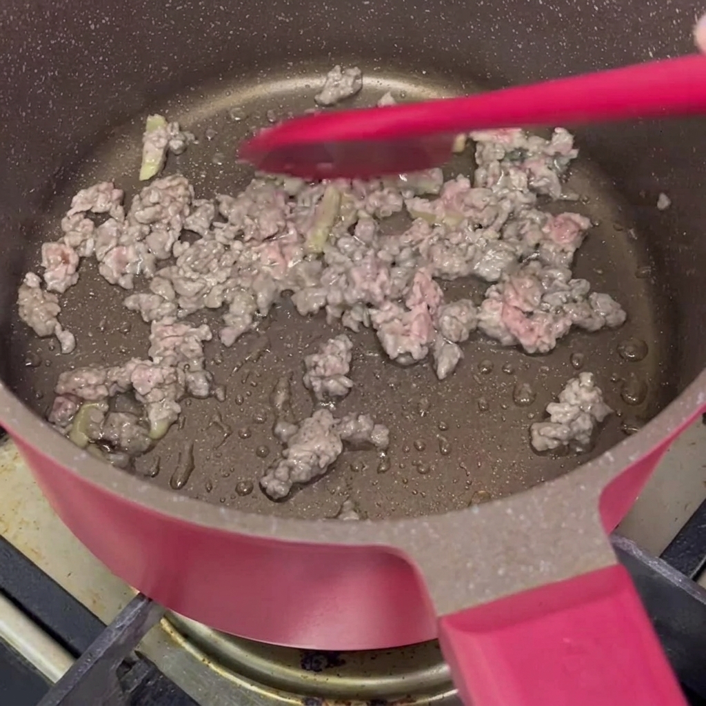 Marinated minced beef being stir-fried with a red silicone spatula in a pink non-stick pot until the meat changes color.