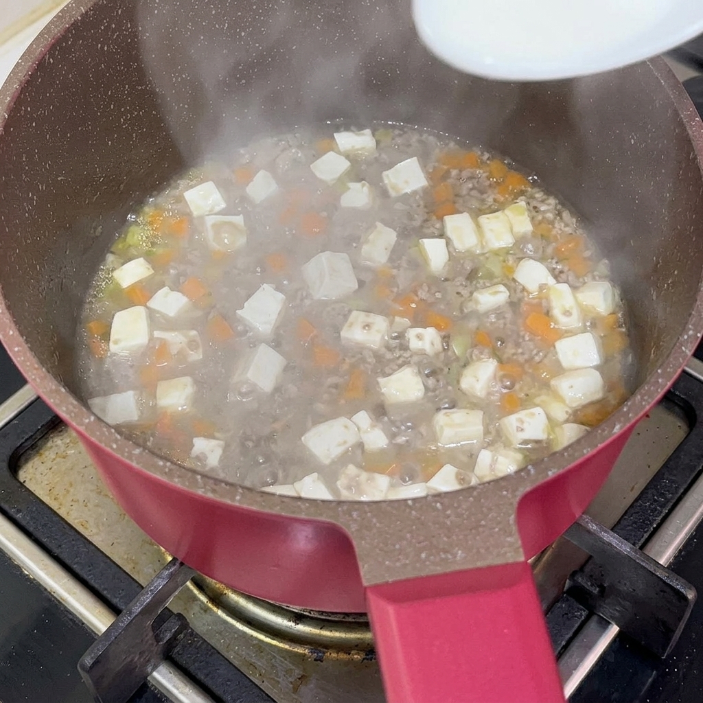 A white spoon pouring translucent starch slurry into a simmering pot of beef, tofu, and carrots to thicken the gravy.