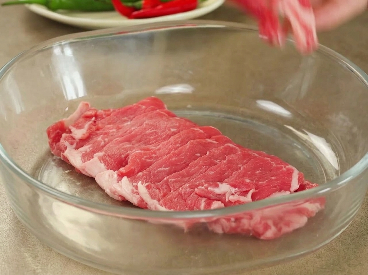 Freshly sliced raw beef arranged neatly inside a large clear glass mixing bowl ready for marination.