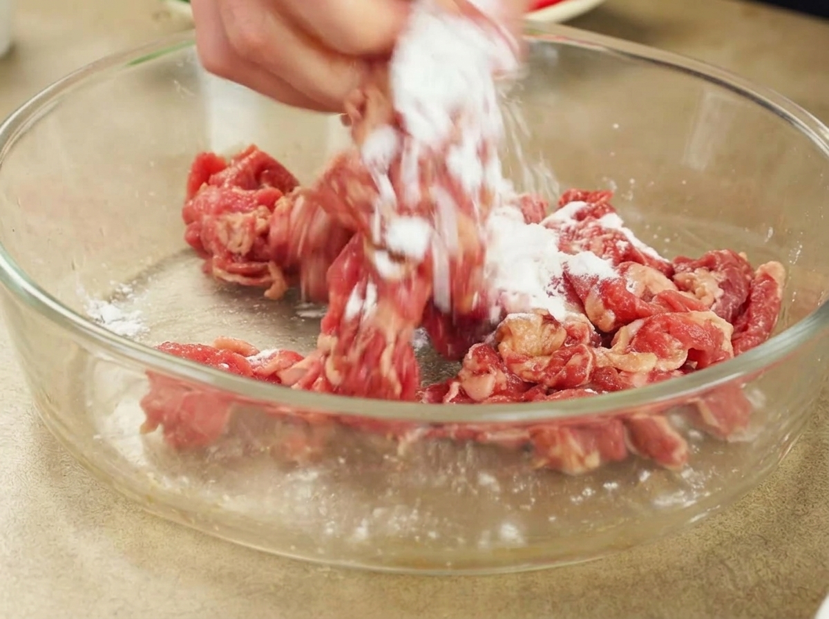 A hand mixing white cornstarch into marinated raw beef slices inside a glass bowl.