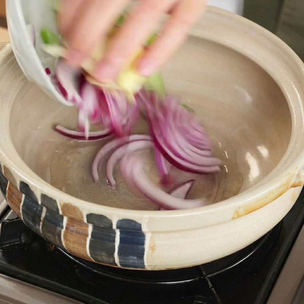 Slices of red onion and other fresh aromatics being dropped into a heated clay cooking pot.