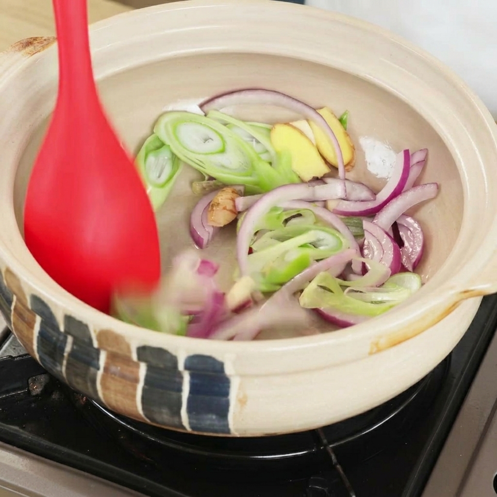 A red spatula stir-frying sliced red onions, scallions, and ginger slices inside a heated clay pot.