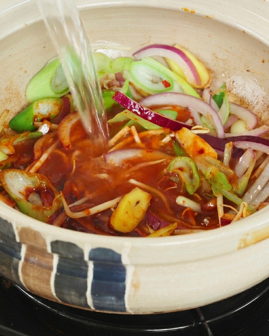A stream of clear boiling water being poured into a ceramic pot filled with onions, scallions, and red chili broth.