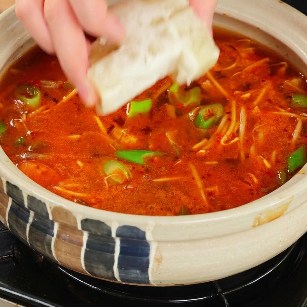 A hand dropping a square, light-colored bouillon cube into a bubbling pot of bright red spicy soup.