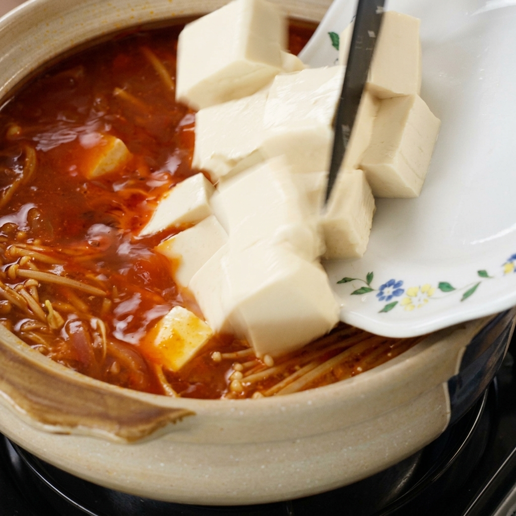 Cubes of fresh white tofu being slid from a floral-patterned plate into a simmering pot of red soup filled with enoki mushrooms.