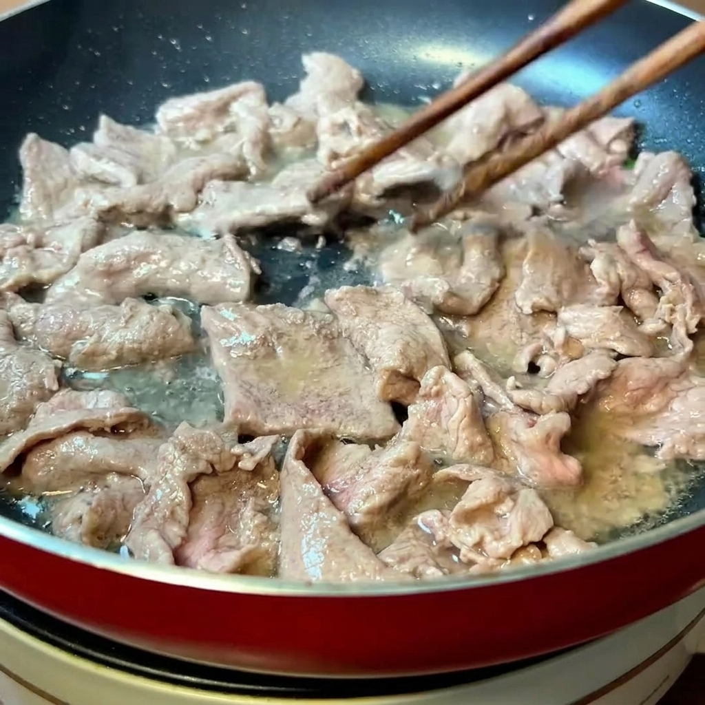 Thinly sliced marinated beef being stir-fried with wooden chopsticks in a heated pan until the surface changes color.
