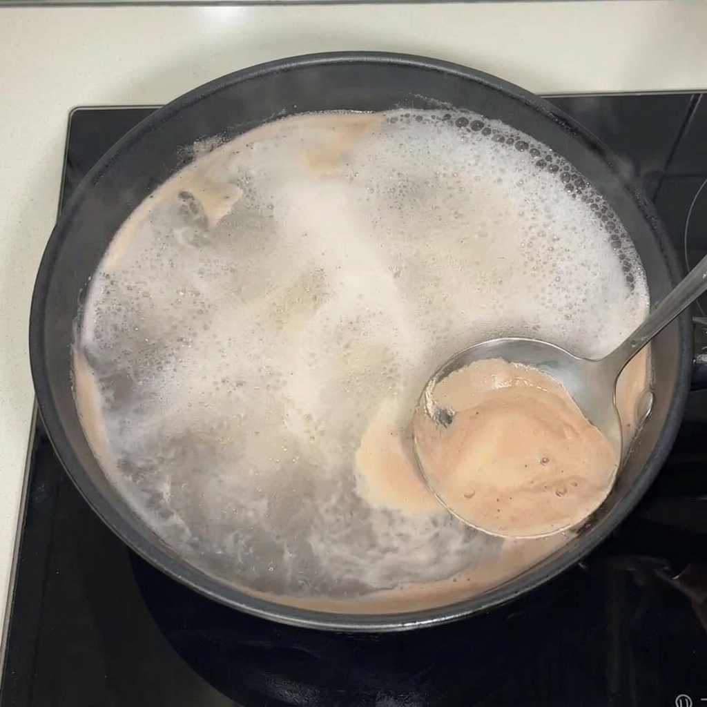 A metal ladle skimming brown foam and impurities from the surface of a boiling broth in a black pan.
