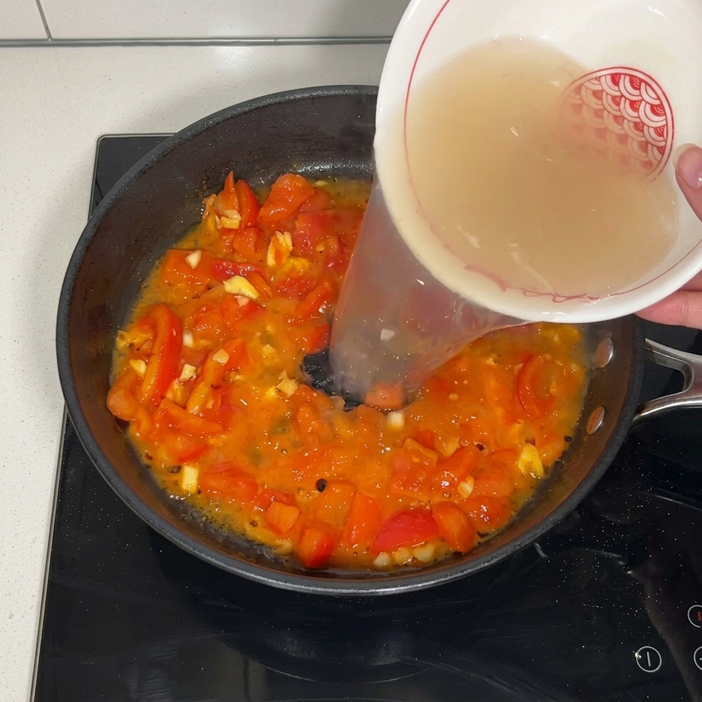 Clear beef broth being poured from a patterned white ceramic bowl into a pan filled with stir-fried tomatoes.