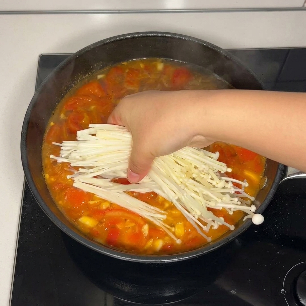 A hand adding a bundle of fresh, white enoki mushrooms into a simmering tomato soup base in a pan.