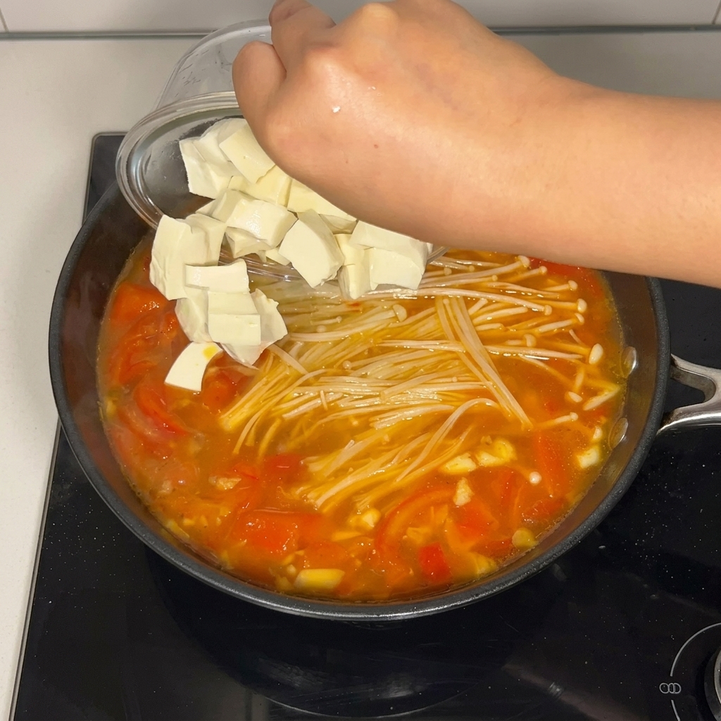 Cubed soft white tofu being poured from a clear glass bowl into a pan of simmering tomato soup with enoki mushrooms.